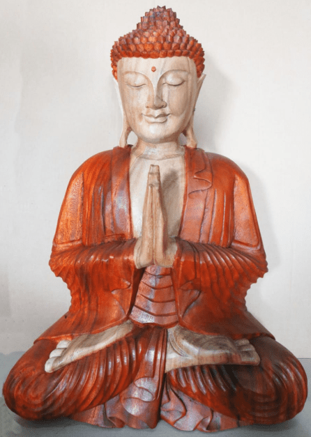 A wooden Buddha statue sits cross-legged, hands in prayer position, with eyes closed, wearing orange robes. The backdrop is plain and subtly lit, emphasizing the statue's detail.