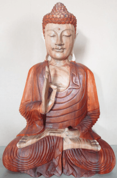 A wooden Buddha statue sits cross-legged in meditation, with one hand raised, the other resting on its lap. The background is a plain wall.
