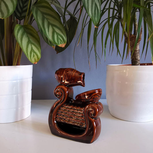A brown ceramic fountain ornament with a spout pours into a basin, placed on a white surface between two large, green-leaved plants in white pots.