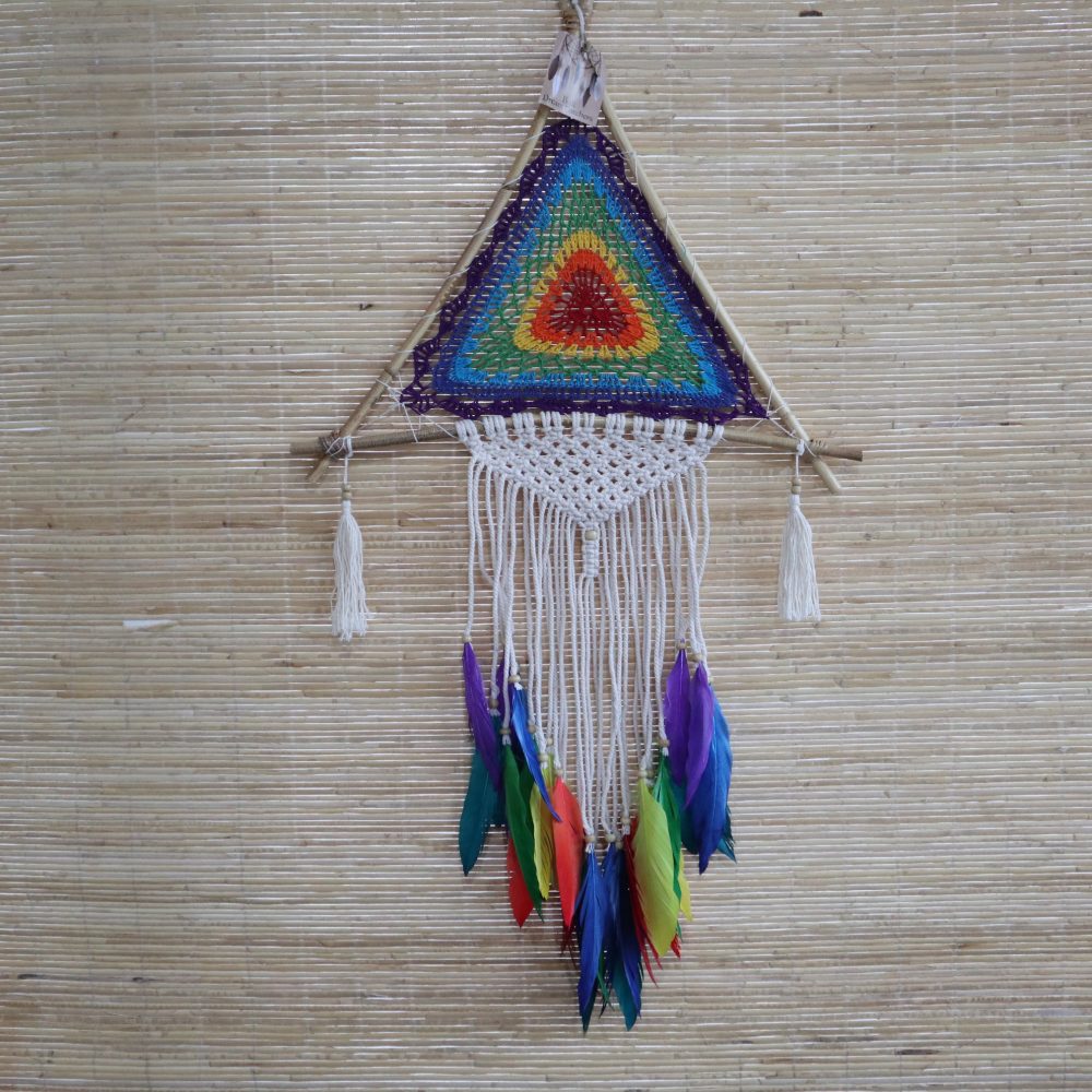 A crocheted rainbow triangle, adorned with feathers and macrame, hangs from a wooden frame against a woven background.