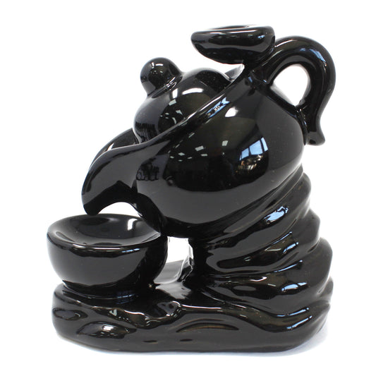 A glossy black ceramic statue depicts a teapot pouring liquid into a bowl, styled to resemble a swirling tornado, on a smooth, reflective surface.