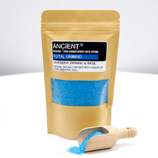 A tan, sealed bag with the text "ANCIENT", "WISDOM / 350G AROMATHERAPY BATH POTION TOTAL UNWIND LAVENDER, ORANGE & BASIL NATURAL SEA SALT INFUSED WITH A BLEND OF PURE ESSENTIAL OILS", filled with blue salt, stands on a white surface near a wooden scoop filled with more blue salt.