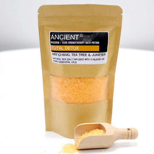 A bag of orange bath salts sits on a white surface with a small wooden scoop. The bag's label reads "ANCIENT WISDOM / 350G AROMATHERAPY BATH POTION TOTAL DETOX MÂY CHANG, TEA TREE & JUNIPER NATURAL SEA SALT INFUSED WITH A BLEND OF PURE ESSENTIAL OILS."