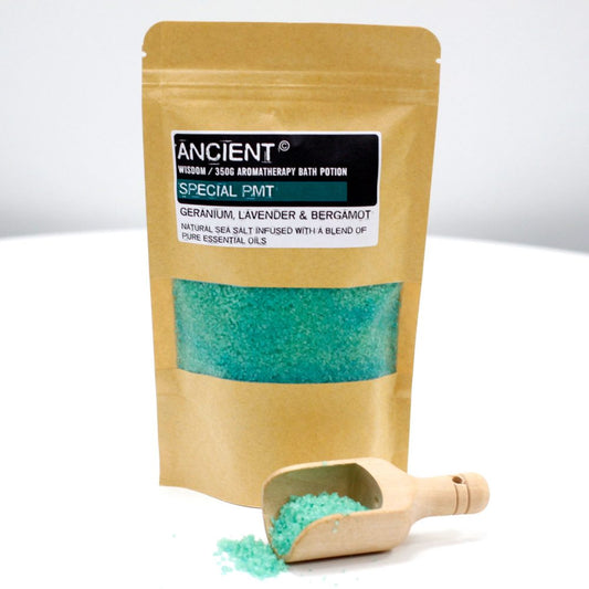 A brown, paper bag of "ANCIENT WISDOM / 350G AROMATHERAPY BATH POTION SPECIAL PMT GERANIUM, LAVENDER & BERGAMOT NATURAL SEA SALT INFUSED WITH A BLEND OF PURE ESSENTIAL OILS" sits next to a wooden scoop filled with turquoise bath salts on a white surface.
