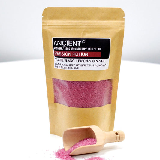 A pouch filled with pink bath salts sits on a white surface, labeled "ANCIENT Wisdom / 350G AROMATHERAPY BATH POTION PASSION POTION, YLANG YLANG, LEMON & ORANGE NATURAL SEA SALT INFUSED WITH A BLEND OF PURE ESSENTIAL OILS". A small wooden scoop spills some of the salts.