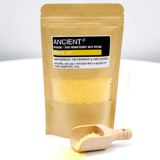 A pouch holds aromatherapy bath potion, visible through a clear window. The pouch is labeled: “ANCIENT, WISDOM / 350G AROMATHERAPY BATH POTION WAKE UP. GRAPEFRUIT, PEPPERMINT & MAY CHANG NATURAL SEA SALT INFUSED WITH A BLEND OF PURE ESSENTIAL OILS". A small scoop rests nearby, spilling potion onto the white surface.