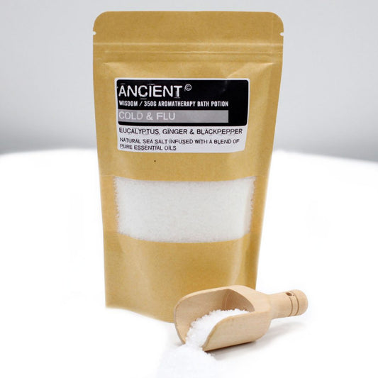 A bag of "ANCIENT WISDOM / 350G AROMATHERAPY BATH POTION COLD & FLU" sits on a reflective white surface with a clear window to show salt, and a small wooden scoop with spilled salt next to it.