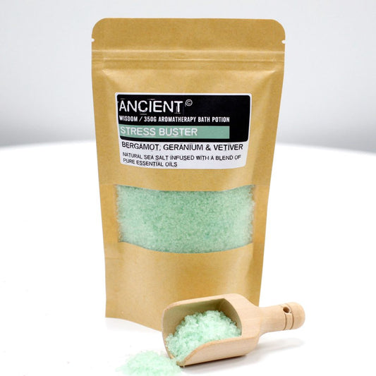 A pouch of bath salts sits with a wooden scoop. The pouch is labeled "ANCIENT Wisdom / 350G AROMATHERAPY BATH POTION STRESS BUSTER BERGAMOT, GERANIUM & VETIVER NATURAL SEA SALT INFUSED WITH A BLEND OF PURE ESSENTIAL OILS" against a white backdrop.