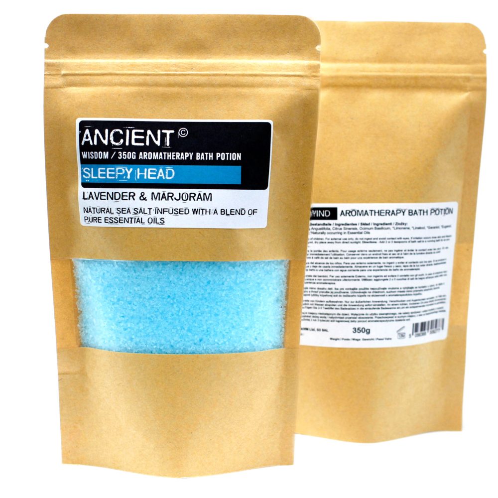 Two stand-up kraft bags contain aromatherapy bath potions. "ANCIENT Sleepy Head: LAVENDER & MARJORAM" bag shows blue salt through a clear window. A partially visible "WIND" aromatherapy bag stands behind it. Bags say "350g."