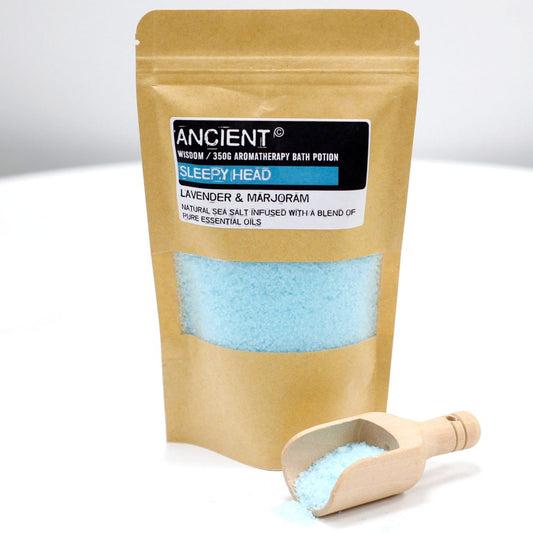 Bath salts fill a kraft paper bag with a window that displays light blue contents. Next to it, a small wooden scoop spills the same salts. The bag reads: "ANCIENT WISDOM / 350G AROMATHERAPY BATH POTION SLEEPY HEAD LAVENDER & MARJORAM NATURAL SEA SALT INFUSED WITH A BLEND OF PURE ESSENTIAL OILS".