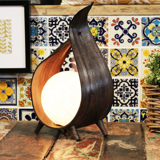 A dark wooden leaf-shaped lamp illuminates a small, round light source. It sits on a rustic wooden table against a colorful tiled wall.