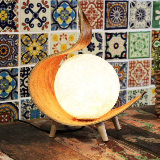 A wooden lamp illuminates a spherical shade; it sits on a rustic table against a colorful tiled wall.