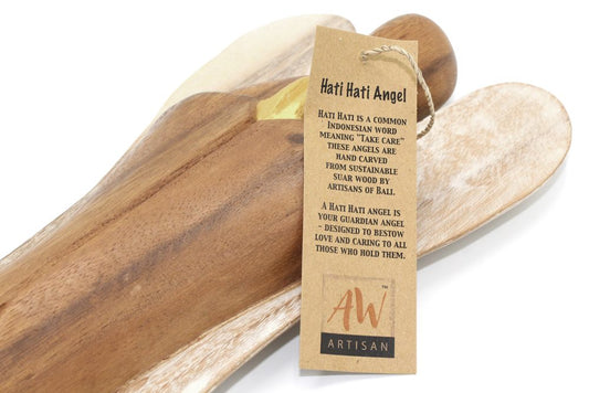 Carved wooden angel rests on pale wooden spoons on a white background; tag reads: Hati Hati Angel Hati Hati is a common Indonesian word meaning "Take Care" These angels are hand carved from sustainable suar wood by artisans of Bali. A Hati Hati angel is your guardian angel - designed to bestow love and caring to all those who hold them. AW™ ARTISAN