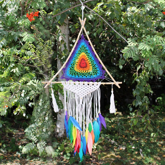 A crocheted rainbow mandala hangs from a wooden triangle, adorned with white macrame and colorful feathers; it's suspended outdoors among lush green foliage.