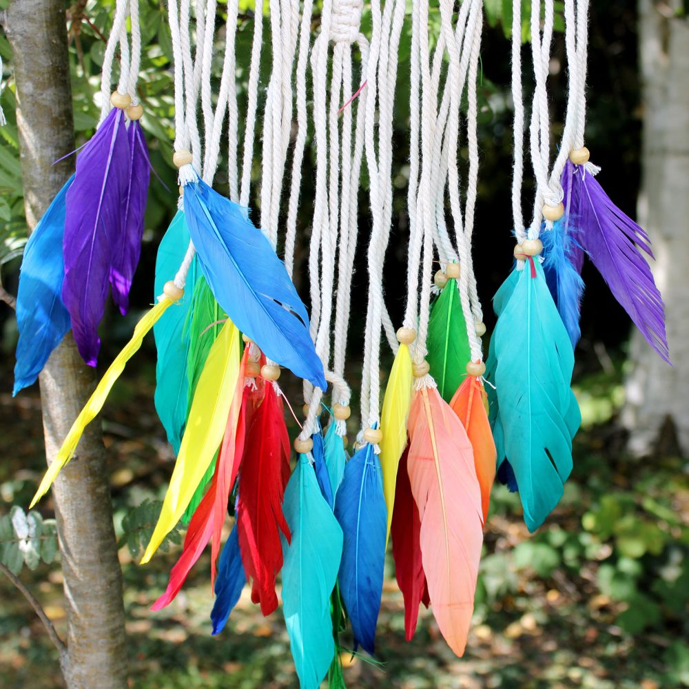 Colorful feathers hang from strings; they are attached to a woven macrame; the setting is outdoors among trees and greenery.