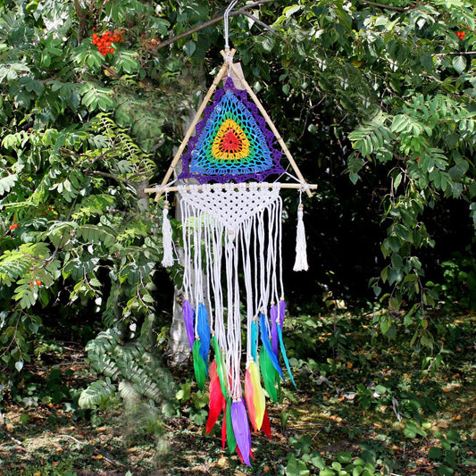 A rainbow-colored crocheted triangle hangs from a wooden frame, adorned with white macrame and colorful feathers. It is suspended in a lush green forest.
