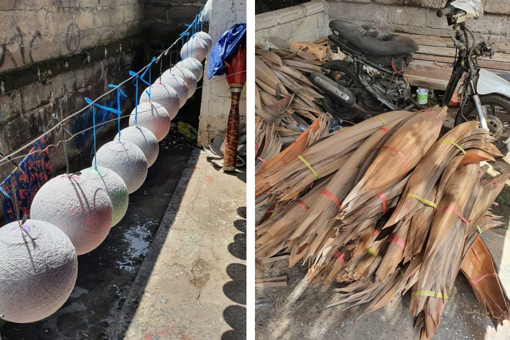 Round lanterns hang drying on strings in an alleyway. Nearby, a pile of palm leaves sits beside a parked motorbike.