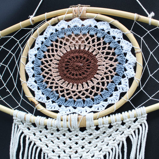 A crocheted mandala hangs from a bamboo hoop, forming a dreamcatcher; off-white macrame fringe hangs below.