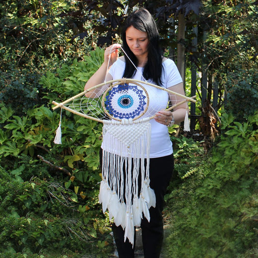 A woman holds a large, eye-shaped dreamcatcher with blue and white crocheted details and white feathers. The dreamcatcher is made of wood and string. She is standing outdoors in a garden.