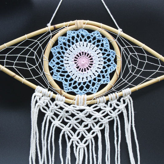 A crocheted mandala hangs within a teardrop-shaped bamboo frame, adorned with macrame fringe; it is suspended against a dark background.