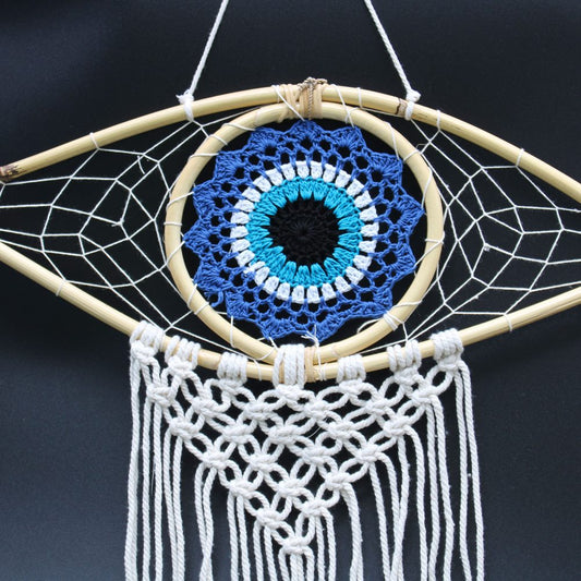 A crocheted evil eye dream catcher hangs against a dark background. The eye is blue, teal, white, and black. Off-white macrame cords form a fringe below.