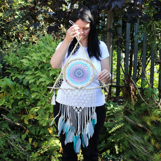 A triangular dreamcatcher, featuring a crocheted mandala and feathers, is being held by a woman. She is adjusting a string in a garden setting.