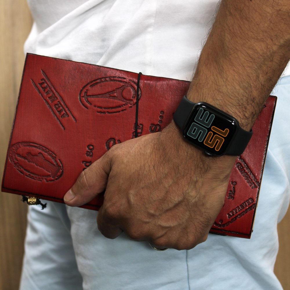 A person's arm holds a red leather journal embossed with "AIR MAIL" and other text, also displaying the time, 3:15, on a black smartwatch. They are wearing a white t-shirt and light-blue pants.