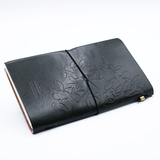 A dark leather journal, held closed by a black elastic band, has “ANCIENT WISDOM” stamped on it and an embossed tree design; it rests on a white background.