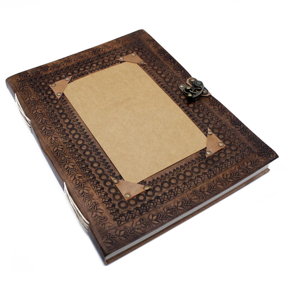 A bound, brown leather journal with an ornate embossed pattern rests on a white surface. A plain rectangular placeholder is mounted on the cover with corner rivets and an antique-looking metal clasp.
