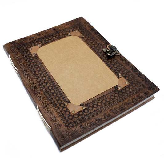 A bound, brown leather journal with an ornate embossed pattern rests on a white surface. A plain rectangular placeholder is mounted on the cover with corner rivets and an antique-looking metal clasp.