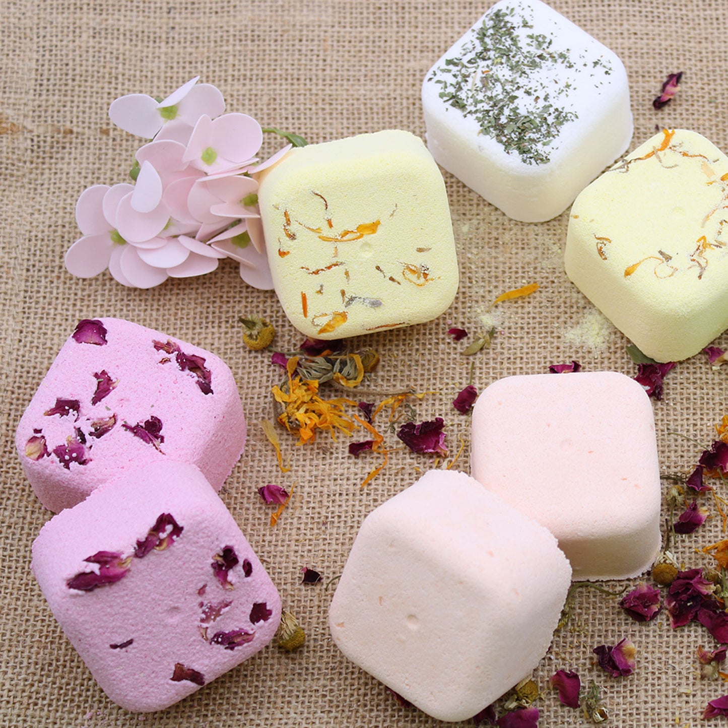 Colorful square bath bombs, adorned with flower petals and herbs, are arranged on a coarse burlap surface, accompanied by pink artificial flowers, creating a rustic, aromatic display.