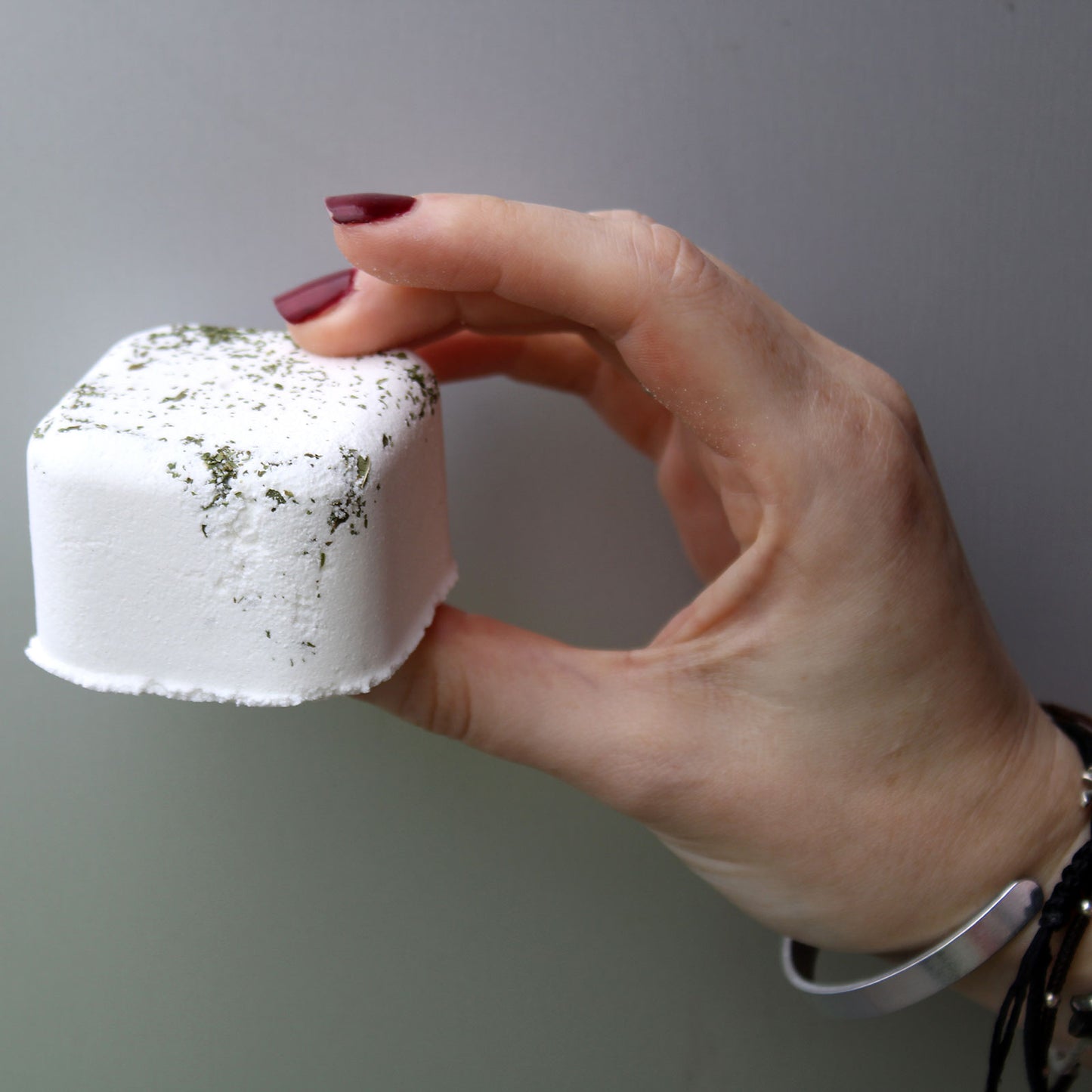 A hand with red-painted nails holds a white, cube-shaped object with green specks, against a plain gray background. The wrist features a silver bracelet and dark wristbands.