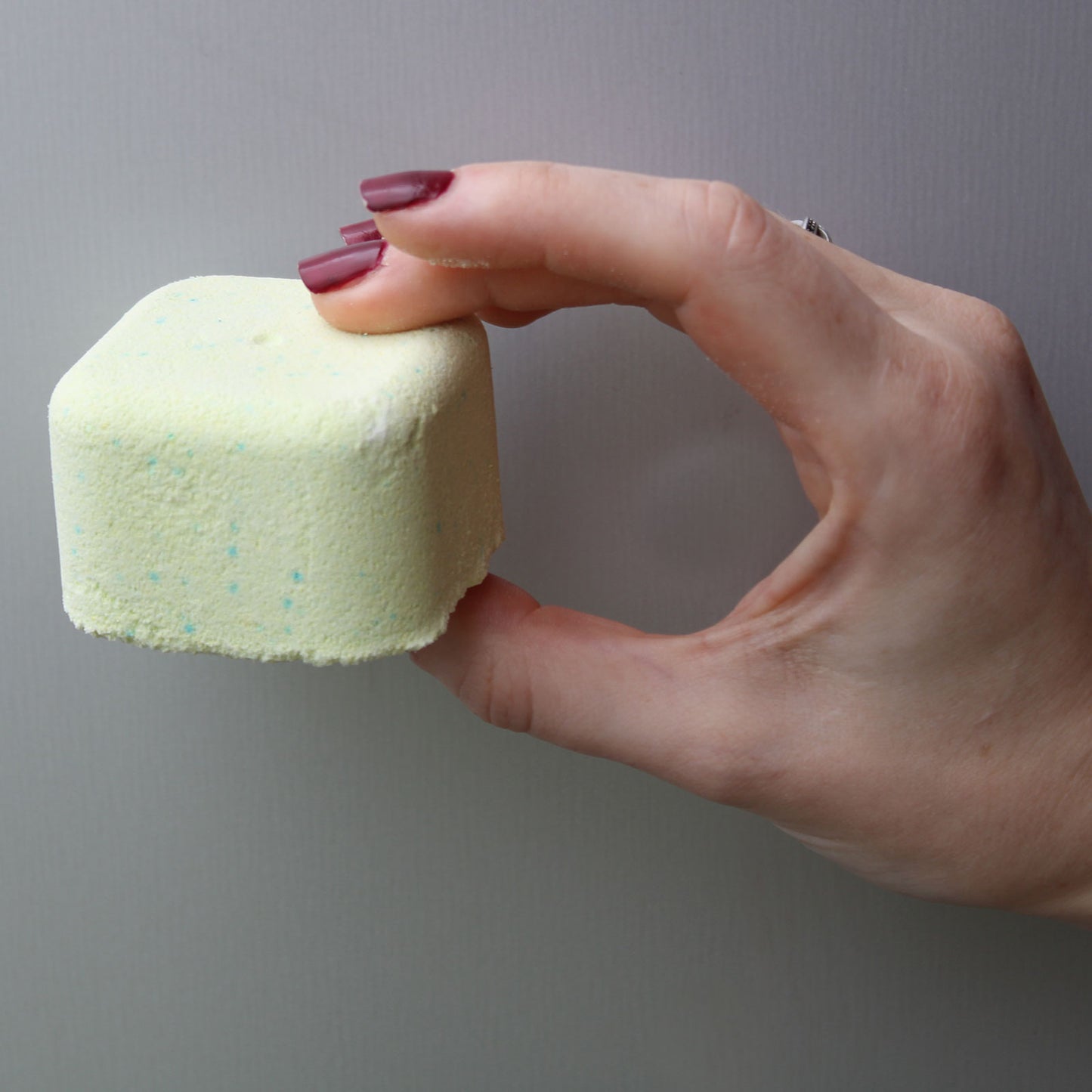 A hand with painted nails holds a light yellow, square-shaped bath bomb, with small blue speckles, against a plain background.