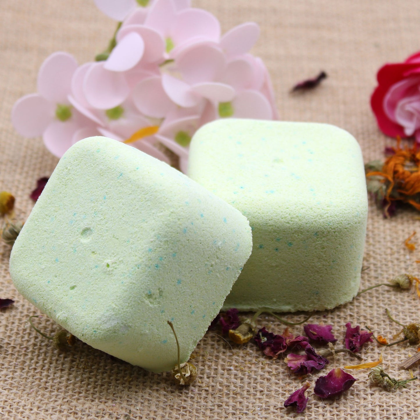 Two light green bath bombs rest on a burlap surface, surrounded by dried flower petals and pink artificial blossoms.