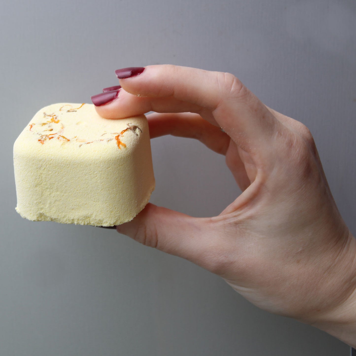 A hand with burgundy nails holds a small, yellow bath bomb with specks, against a plain gray background.