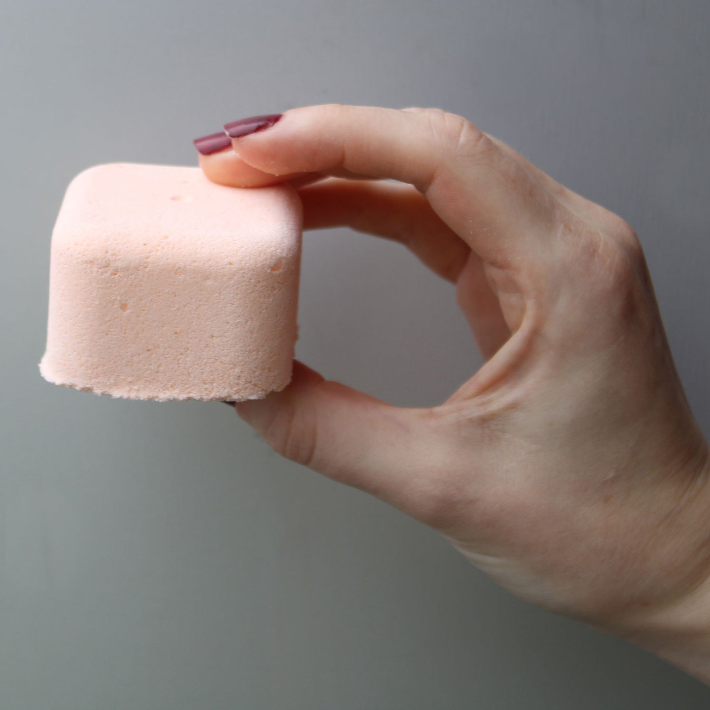 A person's hand holds a pink, square-shaped bath bomb between thumb and forefinger against a plain, light gray background.