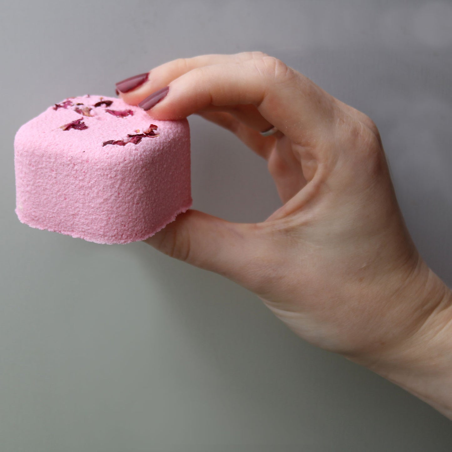 A pink, textured bath bomb with small flower petals is held gently between thumb and forefinger, set against a neutral background.