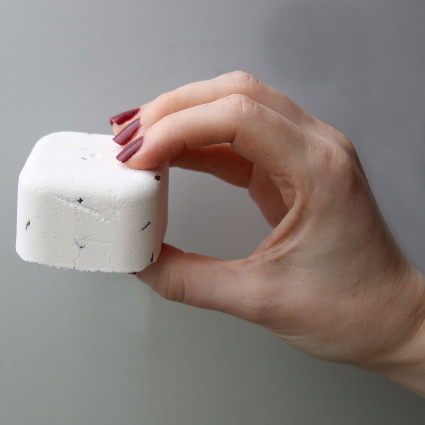 A hand with red nails holds a small, white, rectangular block with black specks against a plain, gray background.