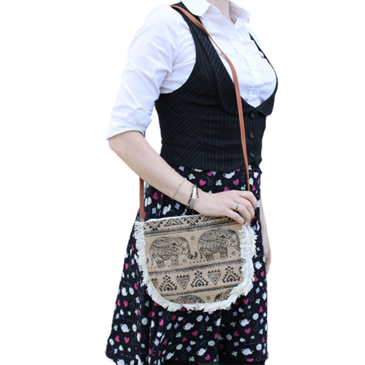 A woman holds an elephant-printed, fringed bag. The bag's strap goes over her shoulder. She wears a white blouse, a black vest, and a skirt patterned with hearts and teacups.