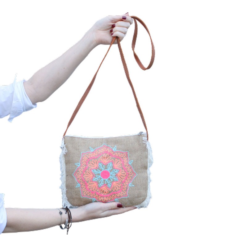 A person holds a burlap purse with a colorful mandala design and a brown strap against a white background. The purse has fringed edges.