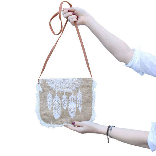 A tan, fringed bag with white feather designs is being held up by two hands against a plain white background. The bag has a brown strap.