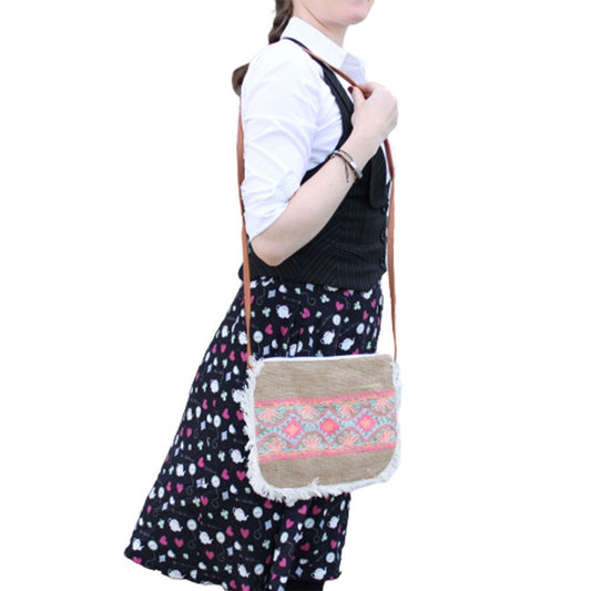 A woman is modeling a small, fringed, woven bag with a strap worn over her shoulder. The woman is wearing a white collared shirt, vest, and a skirt with a print of hearts and teapots.