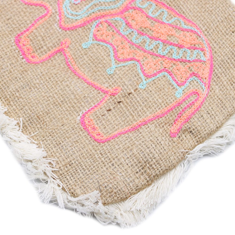 A burlap square with white fringe displays a partial outline of an elephant embroidered in pink, peach, and light blue thread.