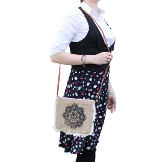 A person holds a brown crossbody bag with a floral mandala design in front. They wear a white blouse, vest, and floral skirt. The background is white.