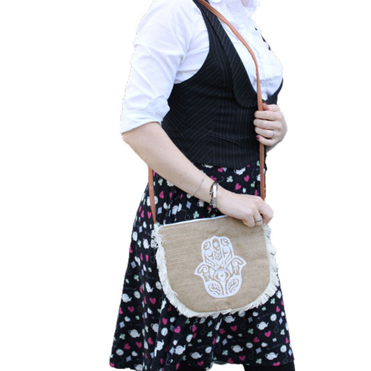 A woman holds a small, tan-colored purse adorned with a white Hamsa hand design and fringed edges. The purse is worn cross-body in front of a heart and spades patterned skirt, white shirt, and black vest.