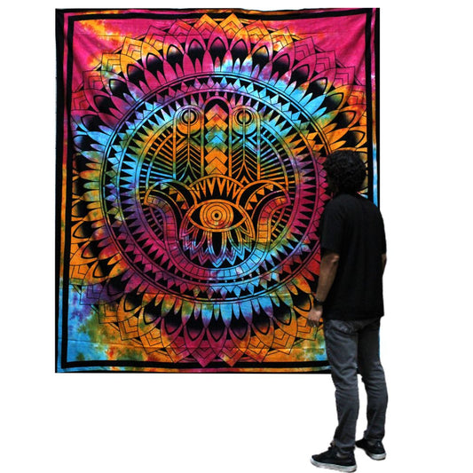 A tie-dye tapestry, featuring a Hamsa hand within a mandala, hangs on a wall; a person observes it.
