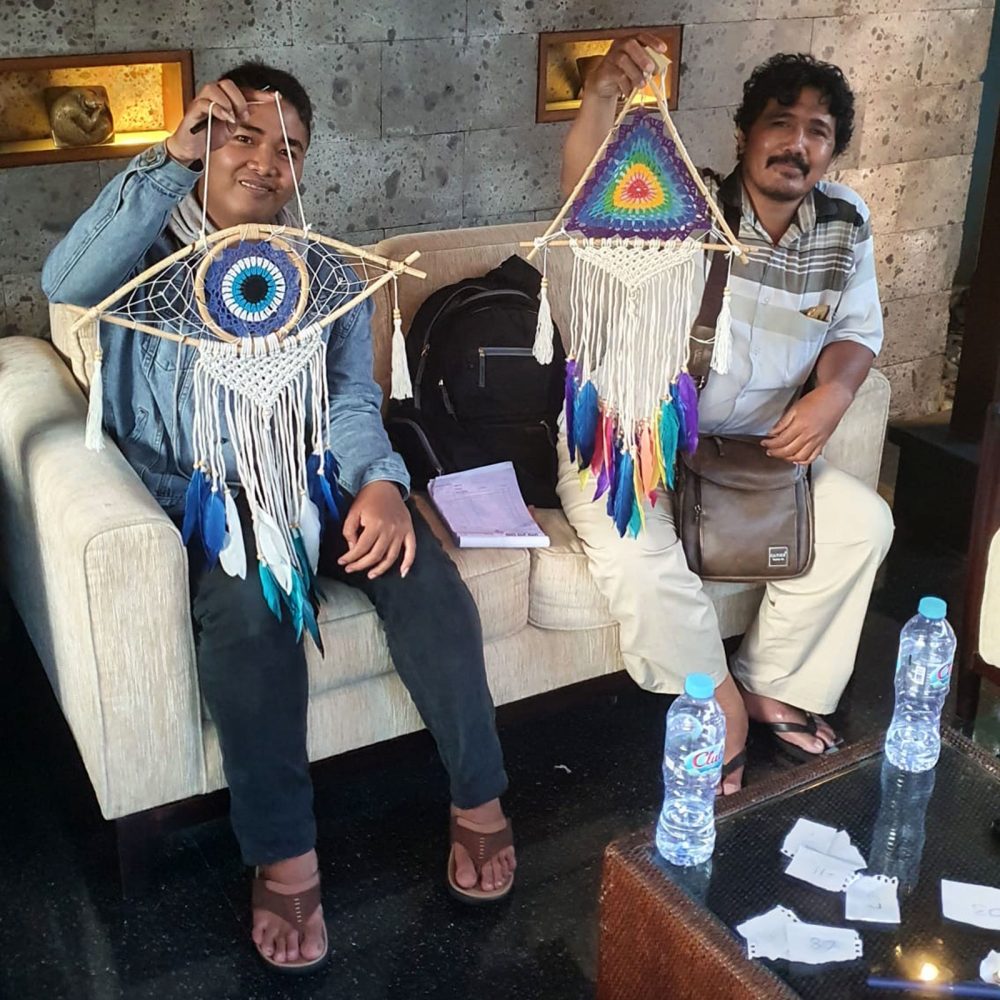 Two men sit on a beige couch, each holding a handcrafted dreamcatcher. One is an eye design, the other a rainbow triangle. They are in a waiting area.