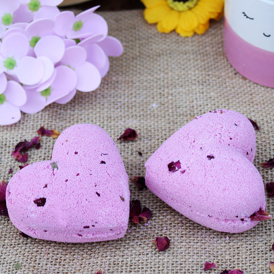 Two heart-shaped pink bath bombs rest on burlap, surrounded by scattered dried rose petals. Nearby, a purple paper flower and a yellow flower add decorative elements to the scene.