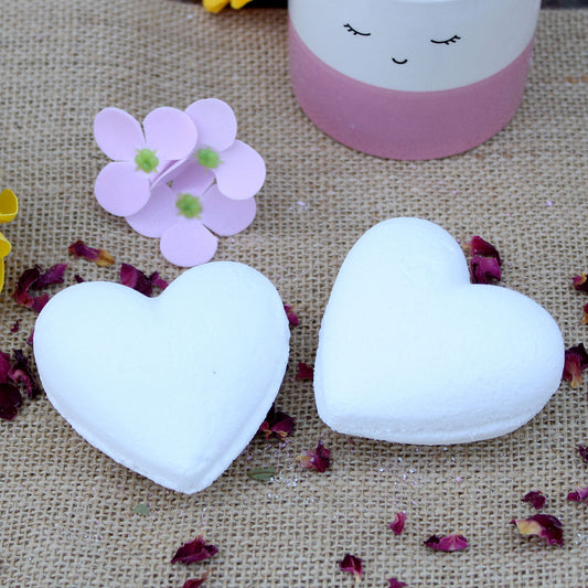 Two white, heart-shaped bath bombs rest on burlap, surrounded by scattered dried flower petals. A cup with a smiling face and pink trim, and paper flowers are nearby.