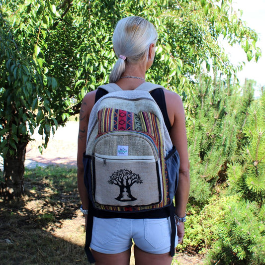 A woman wearing a patterned backpack stands with her back to the viewer. The backpack has a tree with a person meditating at its base. She is in a green outdoor setting.